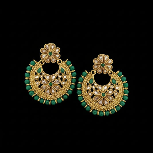 A pair of gold-toned Indian chandbali earrings decorated with green stones and white faux pearls, displayed on a dark reflective surface with the brand logo 'SAHELI MY BEST FRIEND' at the top.