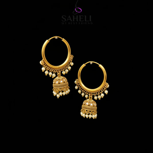 A pair of gold-toned Indian jhumka earrings with small white pearl bead drops, displayed on a black velvet jewelry stand against a gray gradient background. The brand logo 'SAHELI MY BEST FRIEND' is visible at the top.