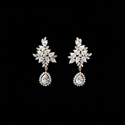 A pair of gold and crystal drop earrings displayed against a solid black background, with the SAHELI brand logo visible at the top center.