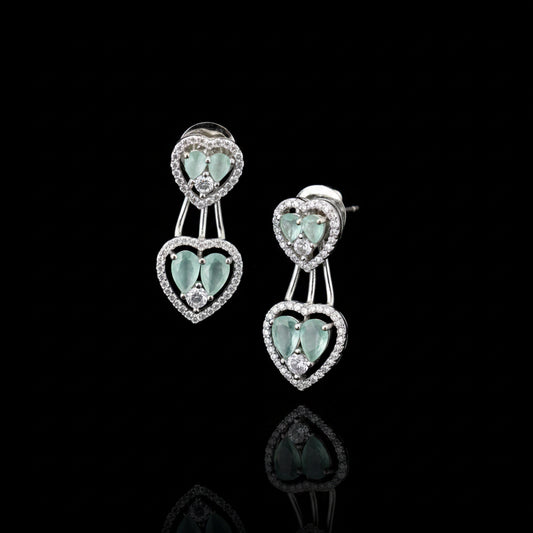 A pair of silver-toned drop earrings with heart-shaped mint green gemstones surrounded by small clear stones, displayed on a gray cushion.