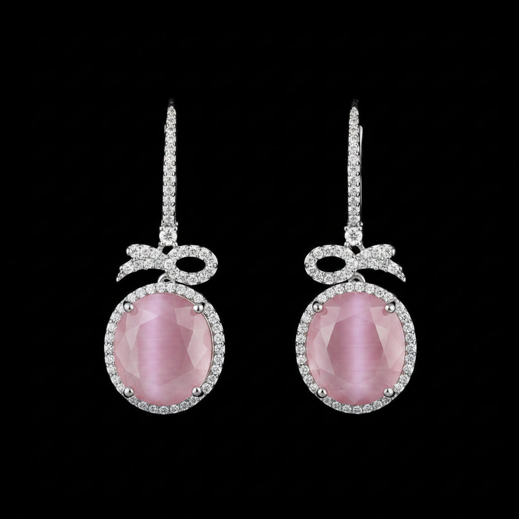 A pair of silver drop earrings with oval pink gemstones and small clear stones, displayed on a textured brown wood background.