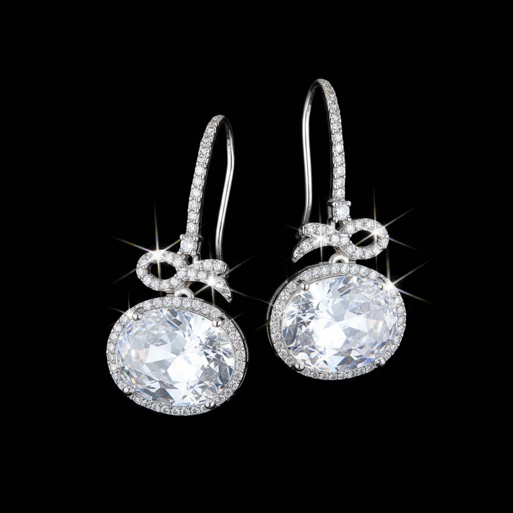 A pair of silver drop earrings with oval clear gemstones and a bow-shaped detail, displayed on a textured brown wood background.