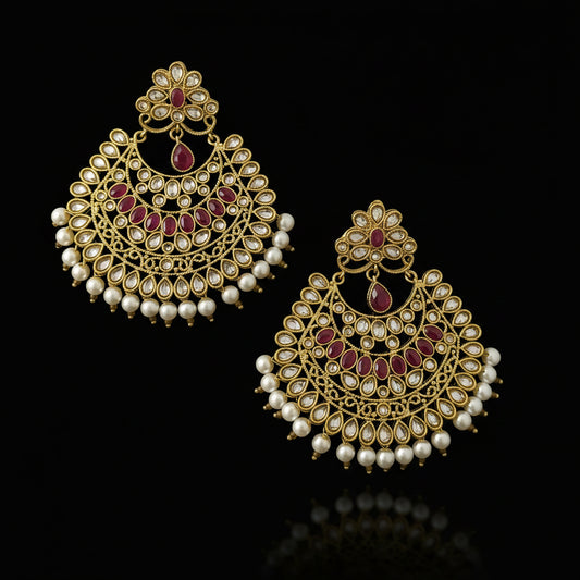 A pair of gold-toned Indian chandbali earrings decorated with white faux pearls, red stones, and clear crystals, displayed on a dark gray reflective surface with the brand logo 'SAHELI MY BEST FRIEND' at the top.
