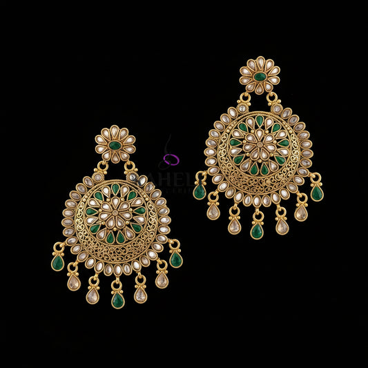 A pair of ornate gold-toned Indian jhumka earrings with green stone accents and clear stones, displayed on a dark gray surface with a Saheli brand logo overlay.