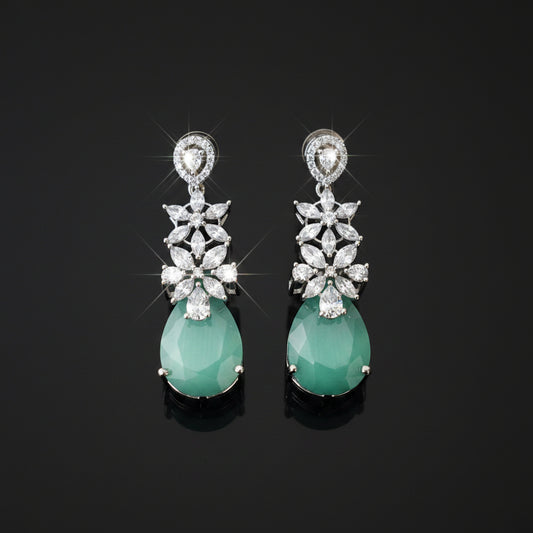 A pair of silver-tone drop earrings featuring a cluster of clear stones in a floral pattern above a teardrop-shaped light green jadeite stone, displayed on a dark reflective surface.