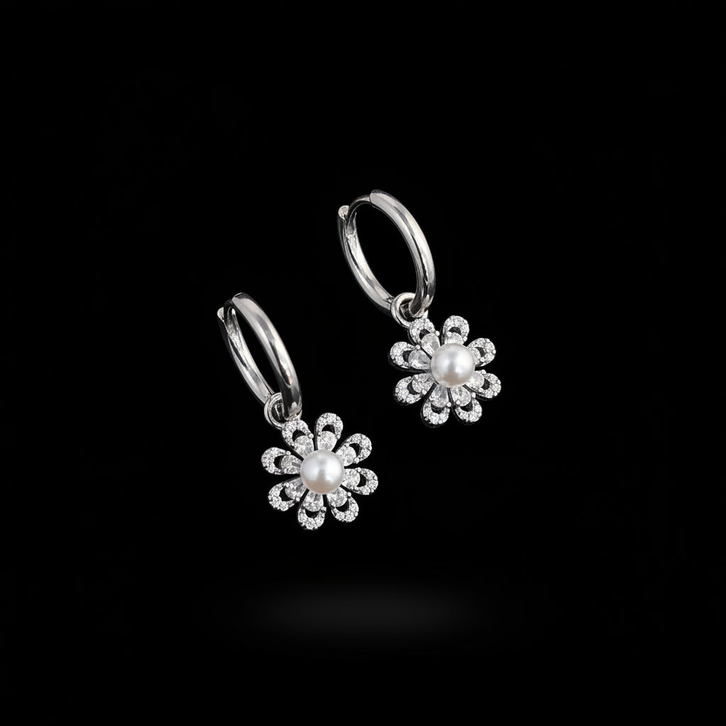 A pair of silver hoop earrings with dangling flower charms, each featuring a central white pearl surrounded by small clear rhinestones, displayed on a white card over a blue background with green leaves in the corner.