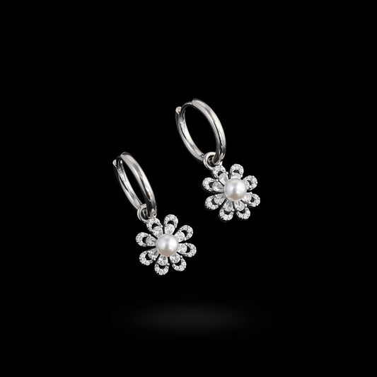 A pair of silver hoop earrings with dangling flower charms, each featuring a central white pearl surrounded by small clear rhinestones, displayed on a white card over a blue background with green leaves in the corner.