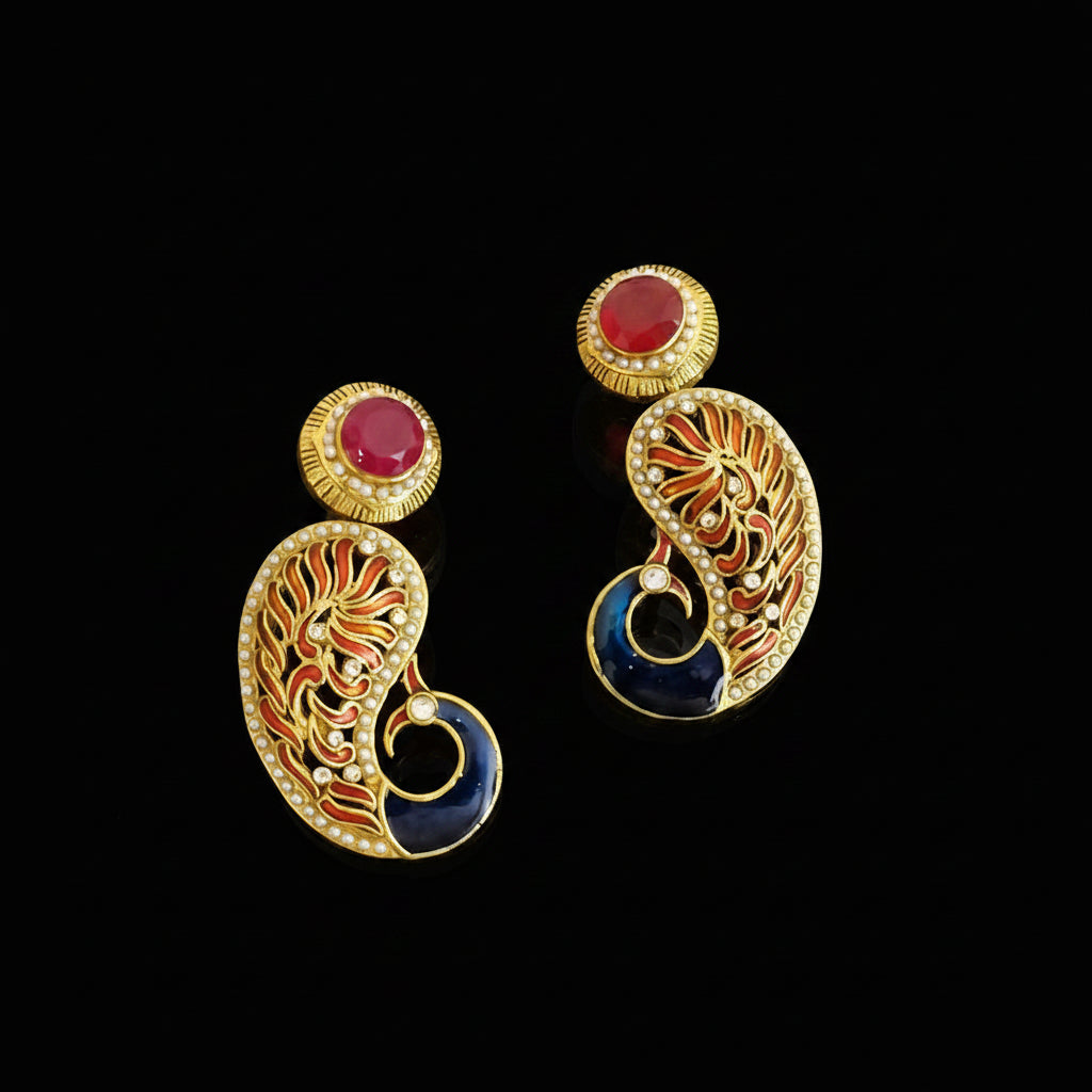 A pair of gold-plated Indian style earrings with red cabochon stones, orange and blue enamel detailing, and small clear stones, displayed on a dark gray reflective surface with the SAHELI MY FANSTRING brand logo at the top.