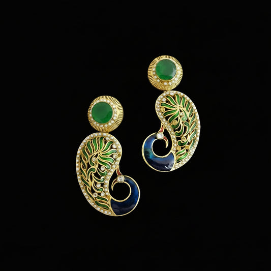 A pair of gold-plated drop earrings with green stone tops, detailed green and gold paisley enameled drops, and a dark blue circular accent, displayed on a dark reflective surface with the SAHeli brand logo above.