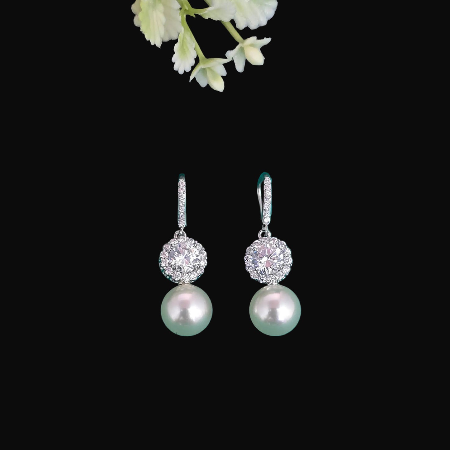 A pair of silver drop earrings with a round cluster of clear pavé stones and a single hanging pearl, displayed on a teal background with decorative stones and flowers.