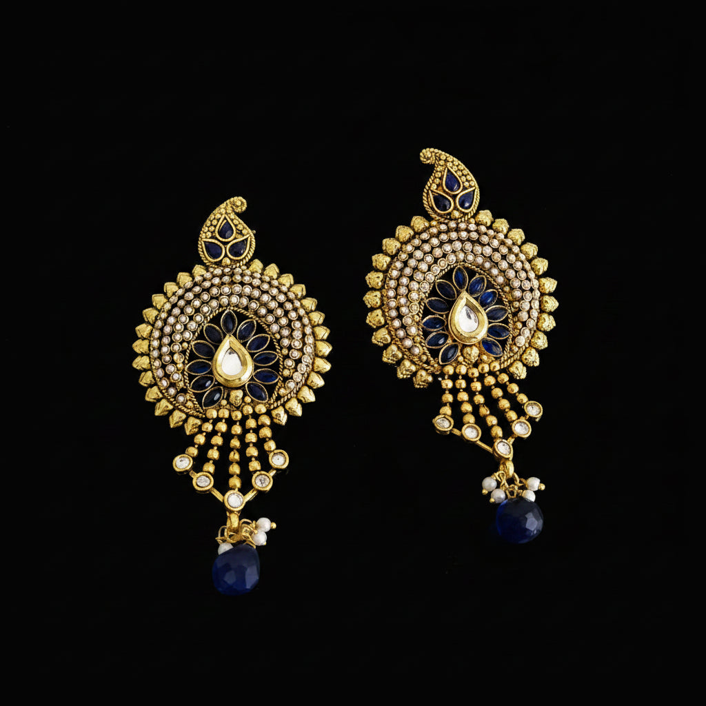 A pair of ornate gold-toned Indian chandbali earrings with blue enamel, white stone accents, and dangling teardrop charms, displayed on a dark gray reflective surface with the Saheli My Best Friend logo at the top.