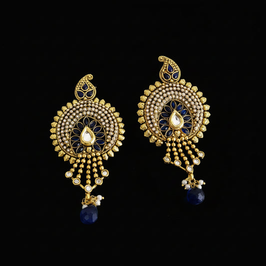 A pair of ornate gold-toned Indian chandbali earrings with blue enamel, white stone accents, and dangling teardrop charms, displayed on a dark gray reflective surface with the Saheli My Best Friend logo at the top.