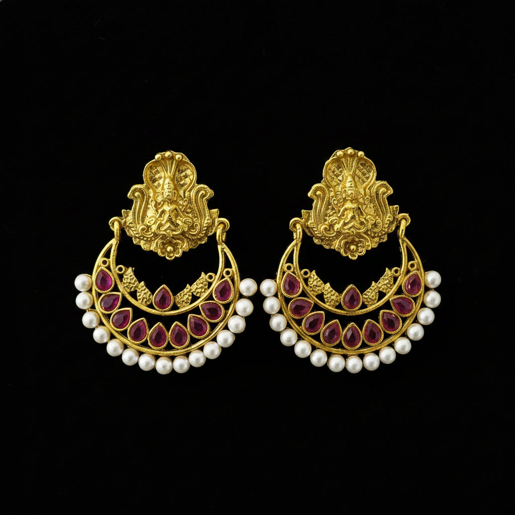 A pair of traditional Indian gold-toned chandbali earrings with red teardrop gemstones and white pearl bead borders, displayed on a dark reflective surface with the SAHeli logo at the top.