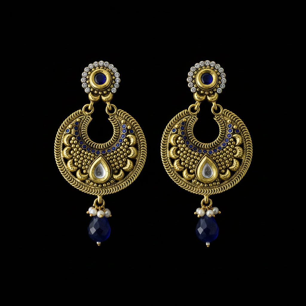 A pair of traditional Indian gold-toned dangle earrings with intricate filigree, white teardrop inlays, small clear stones, and dark blue bead drops, displayed on a dark reflective surface.