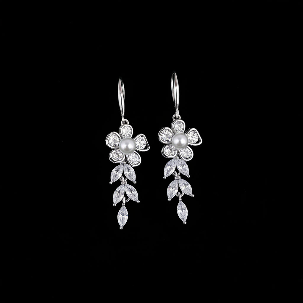 A pair of silver-tone dangling earrings with floral and leaf motifs set with clear rhinestones and a central pearl, displayed on a teal background with decorative stones and greenery in the corner.