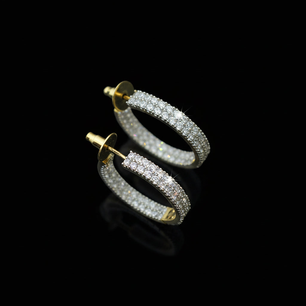 Gold hoop earrings with clear stones on a reflective surface, branded 'SAHELI'.
