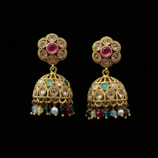 A pair of gold-plated Indian jhumka earrings with multicolor stones and dangling beads, displayed on a solid dark pink background.