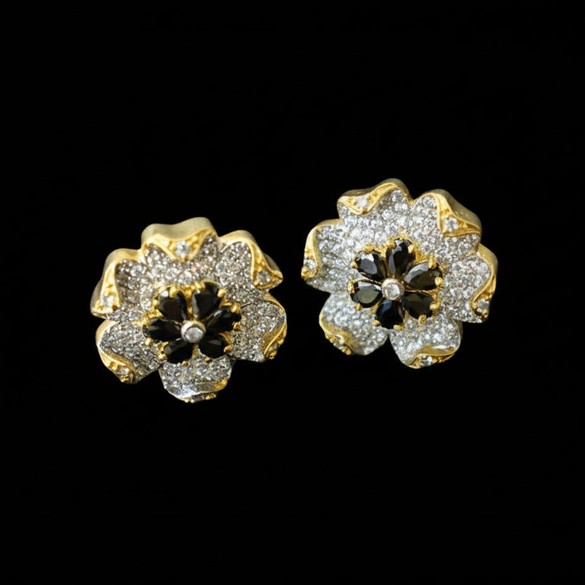Gold and black flower-shaped earrings on a dark background with 'SAHELI' branding.