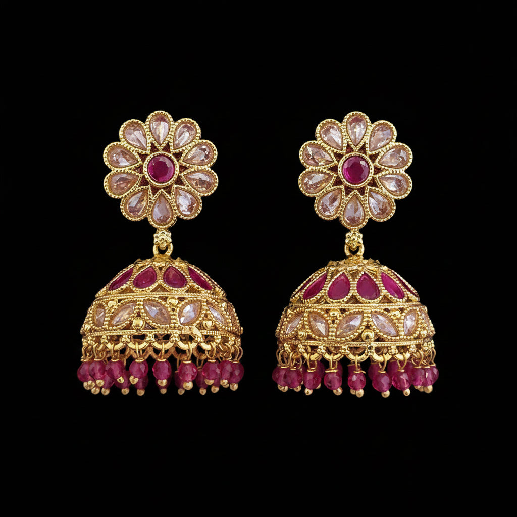 A pair of gold-plated Indian jhumka earrings decorated with pinkish-red stones and clear rhinestones, displayed on a solid dark pink background.