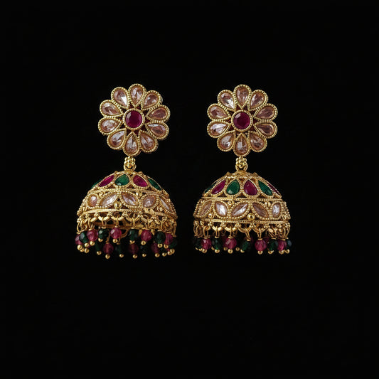 A pair of gold-plated Indian jhumka earrings decorated with clear crystals, pink gemstones, and green gemstones, displayed on a solid dark pink background.