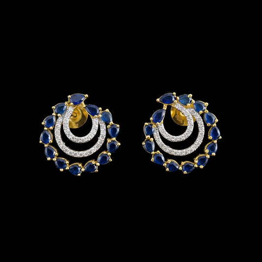 A pair of gold crescent-shaped earrings with blue teardrop stones and clear pavé stones, displayed on a gray reflective surface with the SAHELI MY BEST FRIEND logo above.