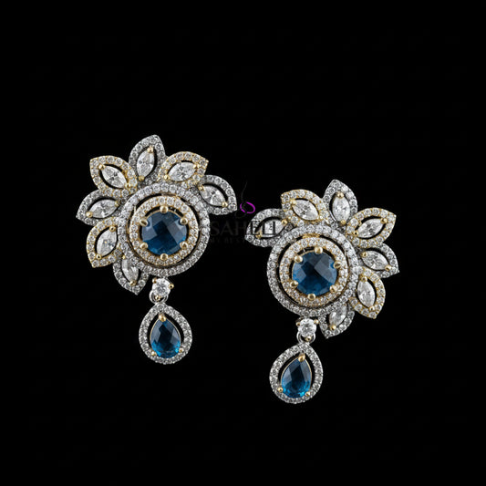 A pair of ornate floral drop earrings displayed on a dark gray stand against a gray background, with the brand name 'SAHELI MY BEST FRIEND' printed above. The earrings feature gold and silver-tone metal settings, clear pavé stones, and deep blue teardrop and round center stones.