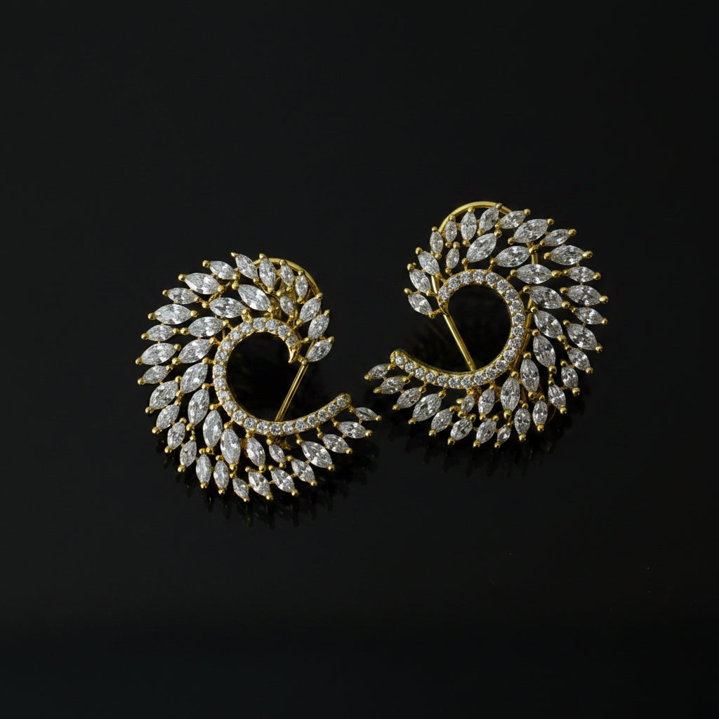 A pair of gold hoop earrings adorned with marquise-cut clear crystals arranged in a layered spiral pattern, displayed on a dark reflective surface with a 'SAHELI MY BEST FRIEND' logo watermark in the upper left.