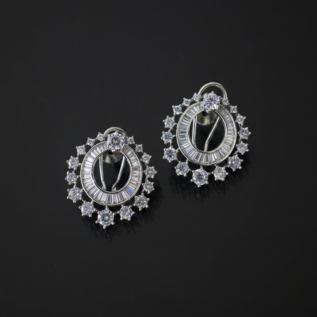 A pair of silver-toned oval hoop earrings adorned with clear baguette and round-cut stones, displayed on a dark reflective surface.