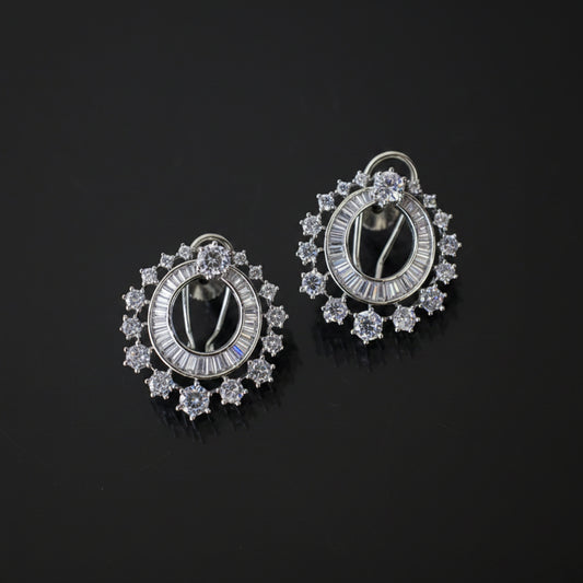 A pair of silver-toned oval hoop earrings adorned with clear baguette and round-cut stones, displayed on a dark reflective surface.