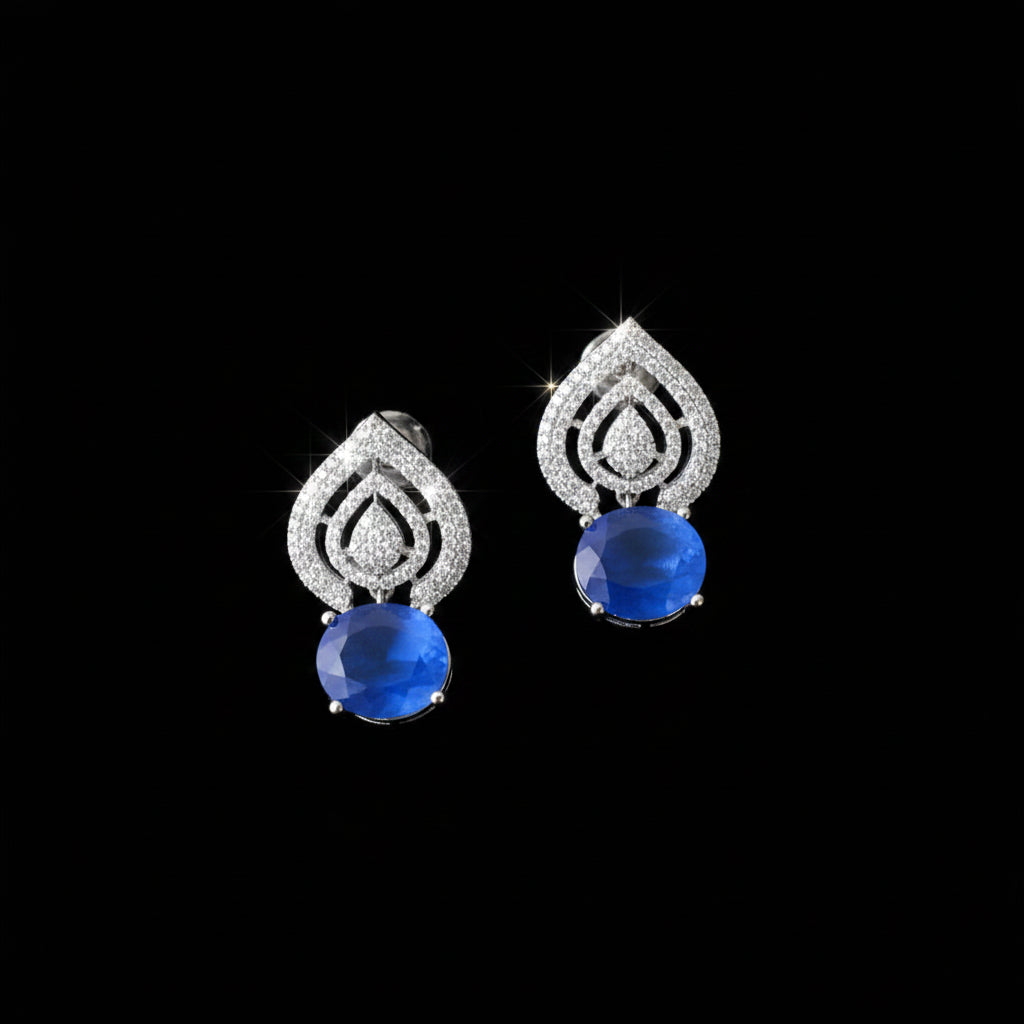 A pair of silver teardrop-shaped earrings with pavé-set clear stones and a round blue cabochon gemstone at the bottom, displayed on a dark reflective surface.