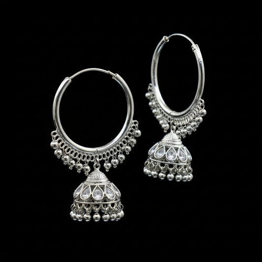 Silver hoop earrings with Jhumka designs on a black stand against a dark background.