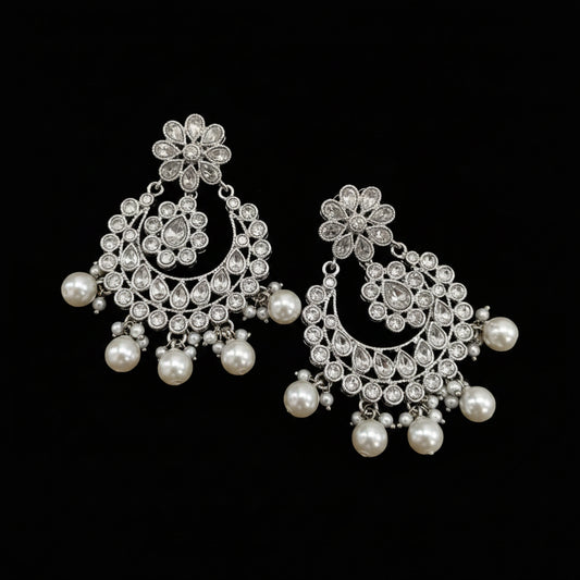 A pair of silver-toned filigree chandelier earrings with white faux pearls, displayed next to a small clear plastic bag containing a matching pair of gold-toned filigree earrings with pearls.