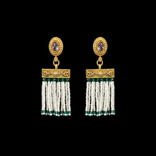 A pair of ornate gold-toned dangle earrings displayed on a dark gray Y-shaped jewelry stand. Each earring features a round gold top with a small oval stone, a rectangular gold connector, and multiple strands of white beads with small green accents.