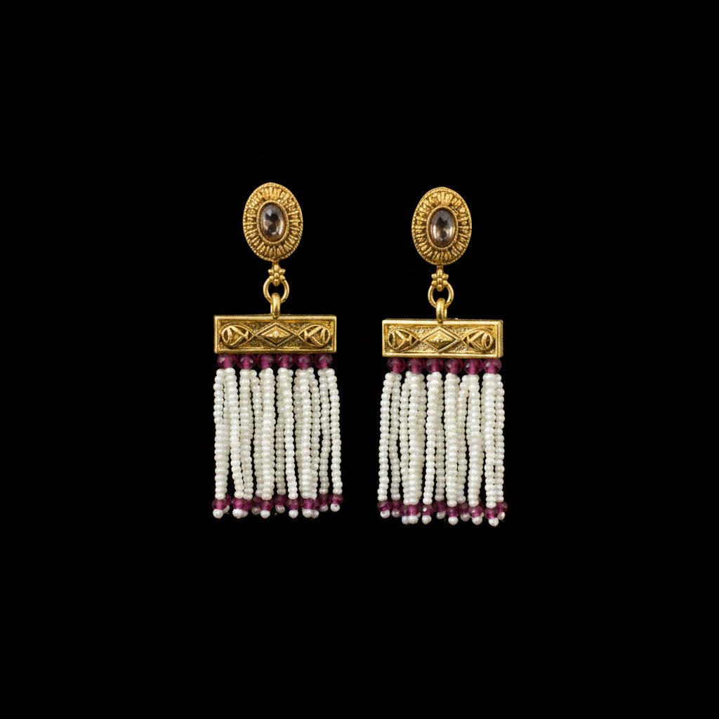 A pair of traditional Indian gold-toned dangle earrings displayed on a black Y-shaped jewelry stand. The earrings feature a circular gold stud with a dark stone, a rectangular gold connector, and multiple strands of white beads accented with small magenta beads at the bottom.