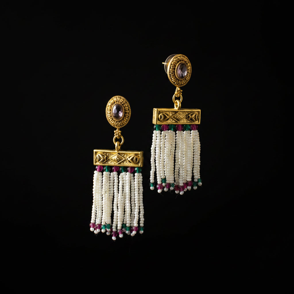 A pair of gold-toned traditional dangle earrings with oval purple stones, ornate gold plates, and white beaded tassels accented with green and pink beads, displayed on a gray jewelry stand.