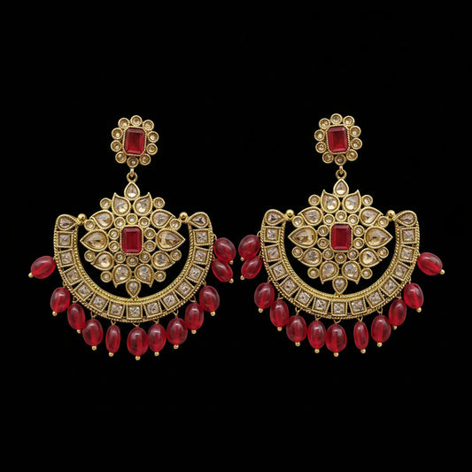 A pair of ornate gold-toned traditional Indian earrings with red stones and dangling red bead chains, displayed on a gray stand against a light gray background. The brand name 'SAHELI MY BEST FRIEND' is visible at the top.
