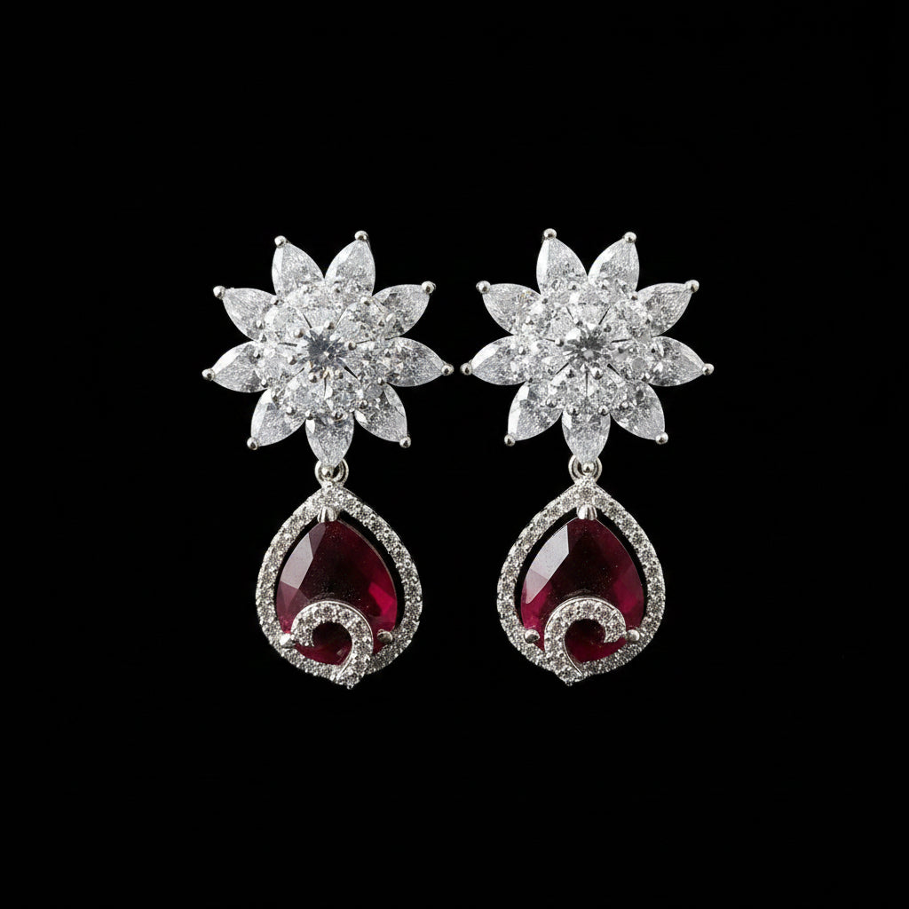 A pair of silver-toned dangle earrings displayed on a black stand against a gray background. Each earring features a flower-shaped top set with clear stones and a teardrop-shaped red gemstone dangle surrounded by small clear stones.