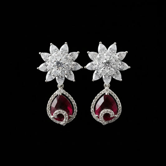 A pair of silver-toned dangle earrings displayed on a black stand against a gray background. Each earring features a flower-shaped top set with clear stones and a teardrop-shaped red gemstone dangle surrounded by small clear stones.