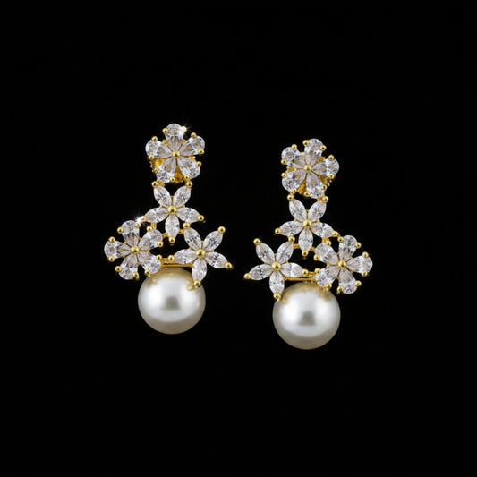 A pair of gold and white pearl drop earrings with clear crystal floral clusters, displayed on a dark gray velvet jewelry stand against a dark gray background.