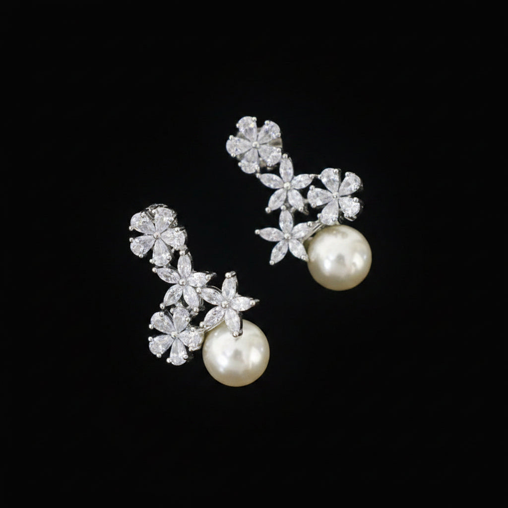 A pair of silver-toned drop earrings with clear floral-shaped crystals and a single white pearl at the bottom, displayed on a gray velvet jewelry cushion against a dark gray background.