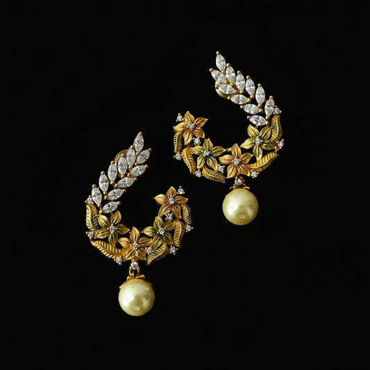 A pair of gold-toned statement earrings with floral and leaf motifs, clear rhinestone accents, and hanging cream-colored pearls, displayed on a dark reflective surface. The brand logo 'SAHELI MY BEST FRIEND' is visible in the top left corner.
