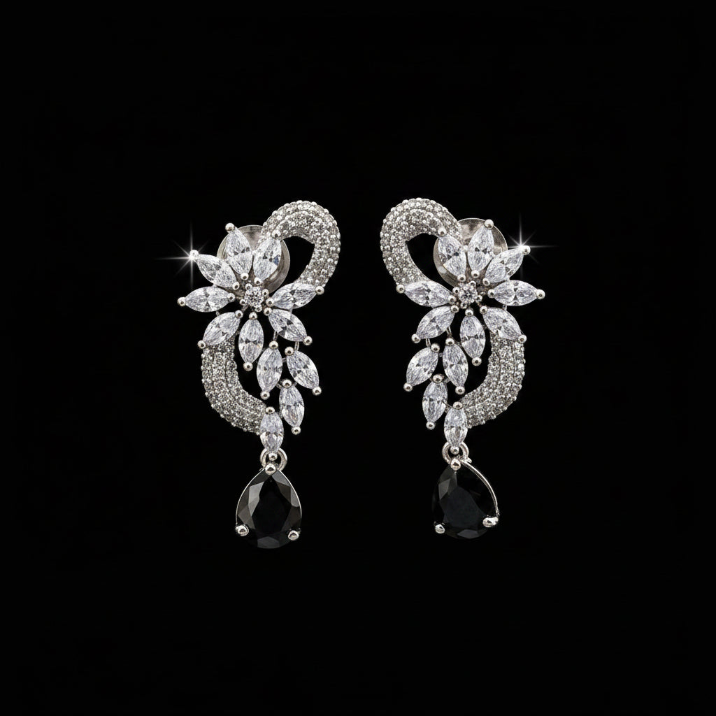 A pair of silver-tone drop earrings with clear marquise and round stones forming a floral heart design, each ending in a black teardrop stone, displayed on a black stand against a gray background. The brand logo 'SAHELI MY BEST FRIEND' is visible in the top left corner.