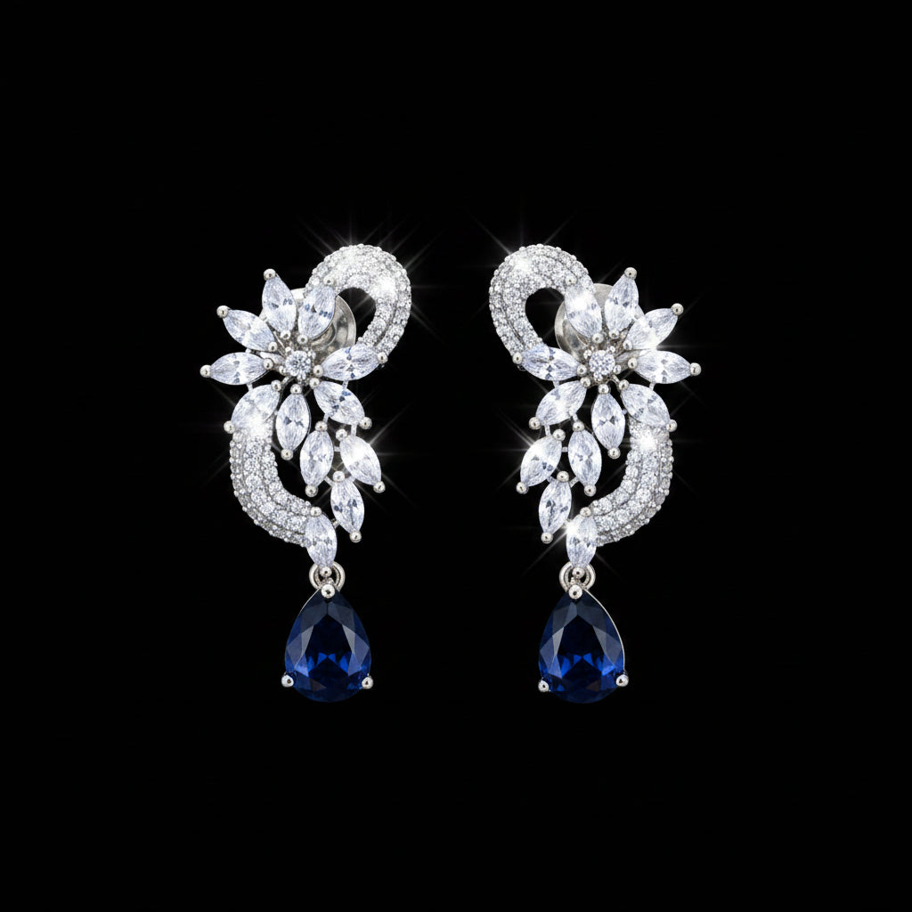 A pair of silver-tone drop earrings with clear marquise and round crystals arranged in a floral scroll motif, each hanging from a teardrop-shaped dark navy blue stone, displayed on a black stand against a gray background. The brand logo 'SAHELI MY BEST FRIEND' is visible in the top left corner.