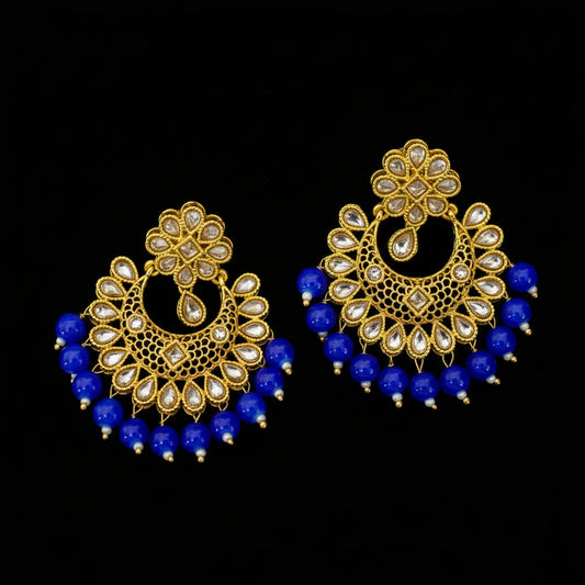 A pair of ornate gold-toned traditional Indian earrings with clear stone embellishments and dangling royal blue bead fringes, displayed on a dark gray surface. The brand logo 'SAHeli' is visible in the top left corner.