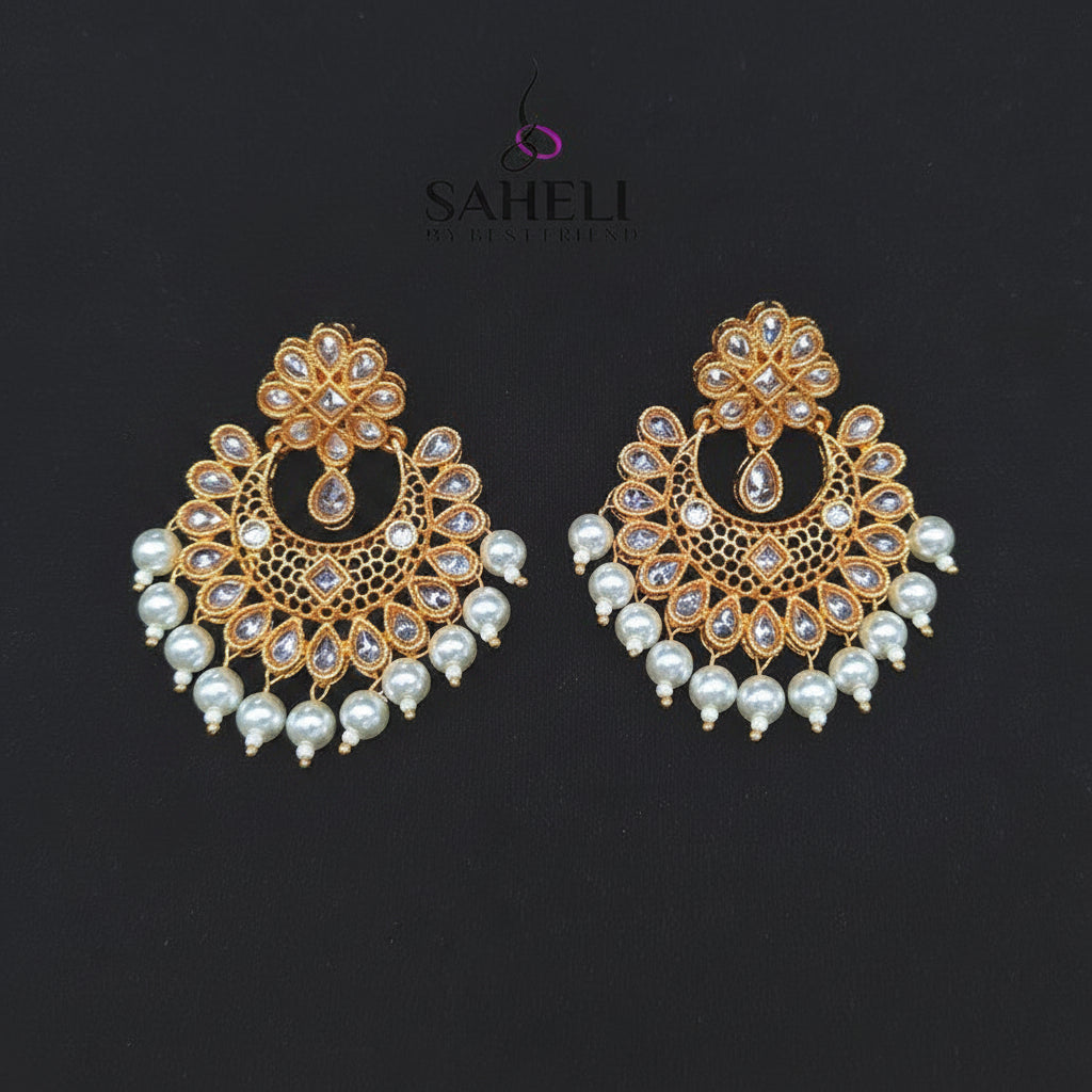 A pair of gold-toned traditional Indian chandbali earrings decorated with white faux pearls and clear stones, displayed on a black background with the SAHeli brand logo above.
