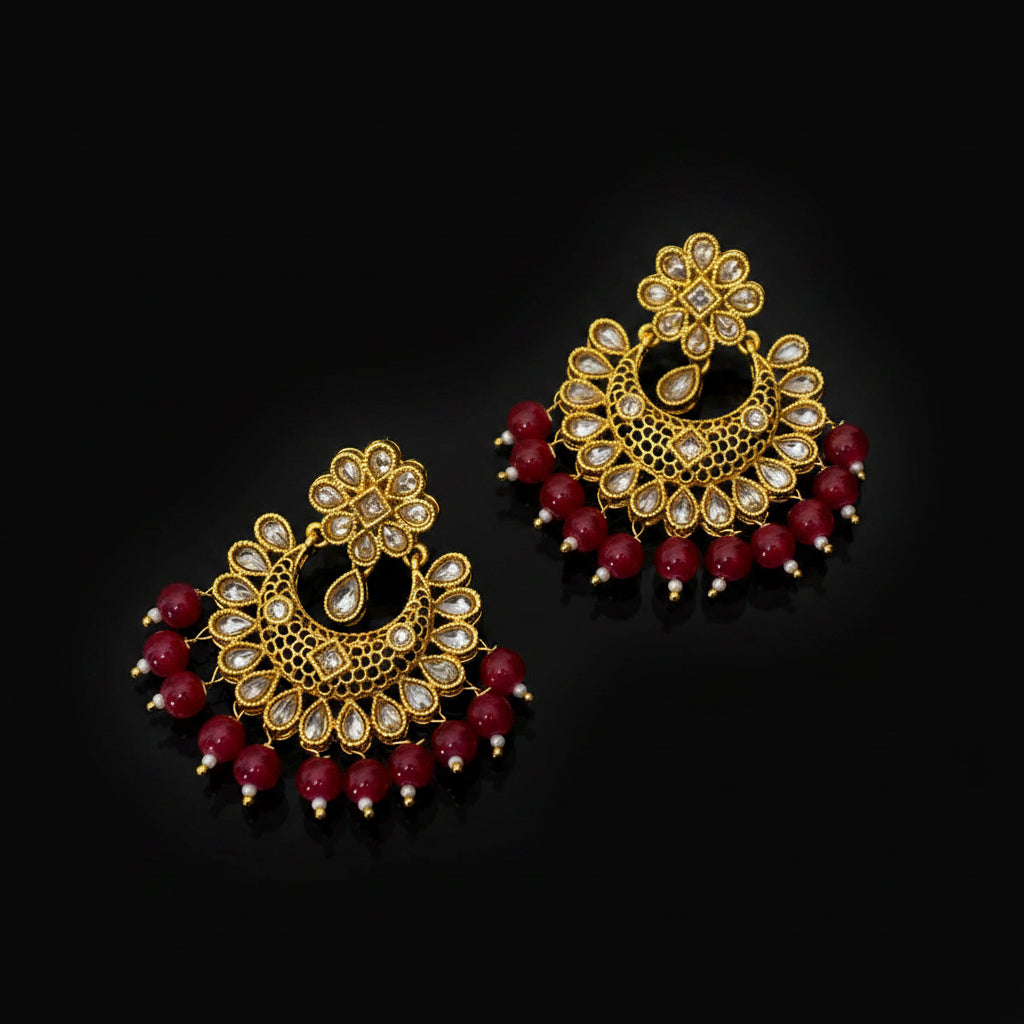 A pair of ornate gold-toned Indian jhumka earrings decorated with clear stones and dangling red beads, displayed on a dark gray reflective surface. A brand tag and the 'SAHeli' logo are visible.