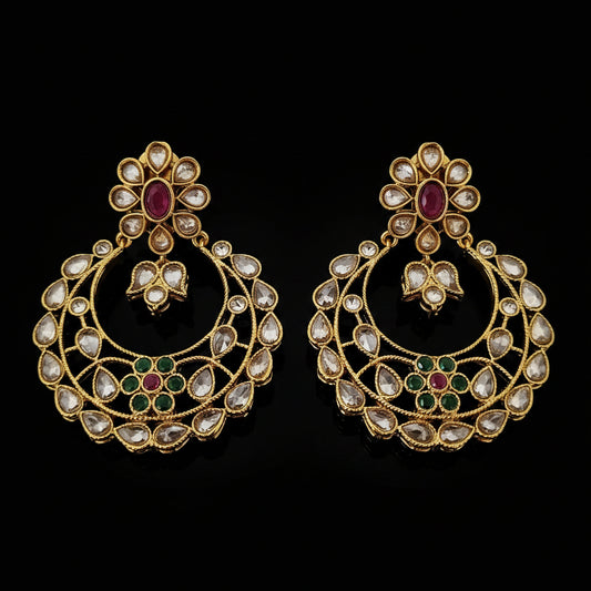 A pair of gold-toned Indian chandbali earrings adorned with clear stones and small red and green enamel accents, displayed on a dark reflective surface with the brand logo 'SAHELI MY BEST FRIEND' above.
