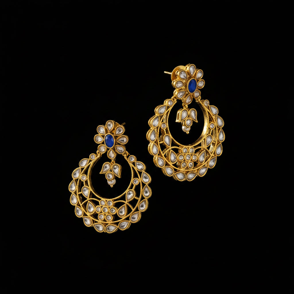 A pair of gold-toned traditional Indian chandbali earrings embellished with white stones and a small blue stone, displayed on a dark gray reflective surface.