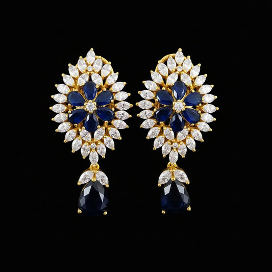 A pair of gold floral drop earrings with deep blue oval stones, surrounded by clear marquise and round crystals, displayed on a black stand against a gray background with the SAHELI MY BEST FRIEND logo at the top.