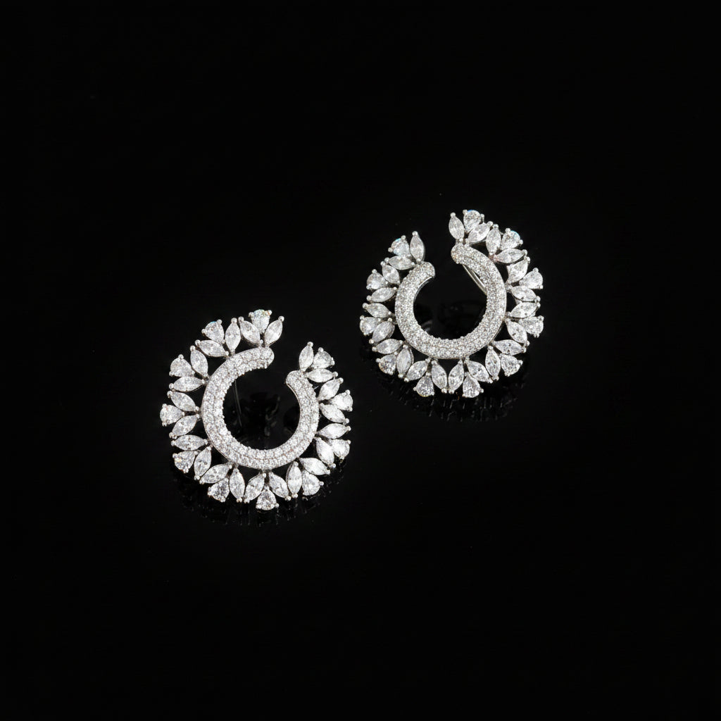 A pair of silver crescent-shaped hoop earrings adorned with sparkling clear stones, displayed on a dark reflective surface with the SAHeli My Best Friend logo in the upper center.